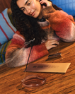 A woman in a fuzzy multicolor sweater leans over a wooden table, with DedCool Incense + Holder set in Mocha Magic burning in front of her